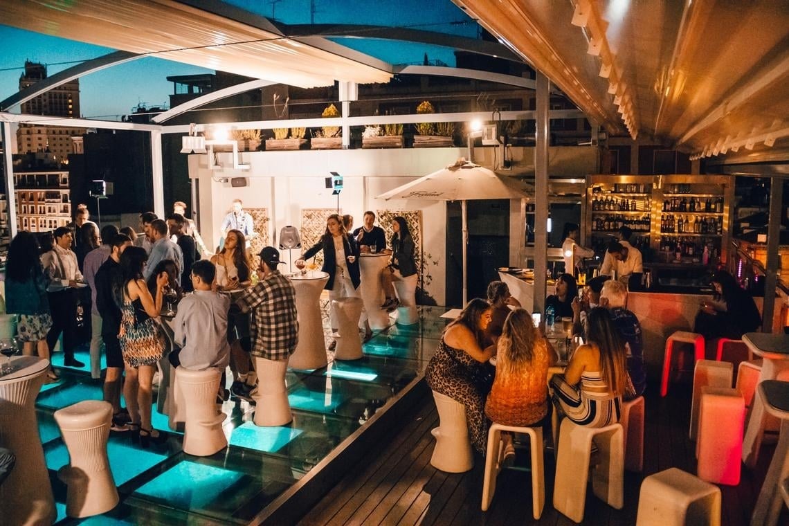 Una terraza de bar con vista urbana al atardecer, donde grupos disfrutan de bebidas y conversaciones. Iluminación cálida,